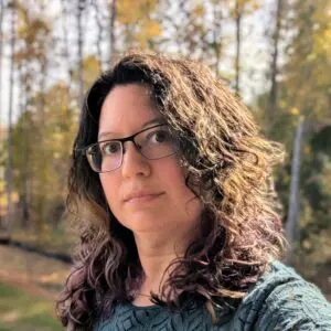 A woman with glasses is standing in front of trees.